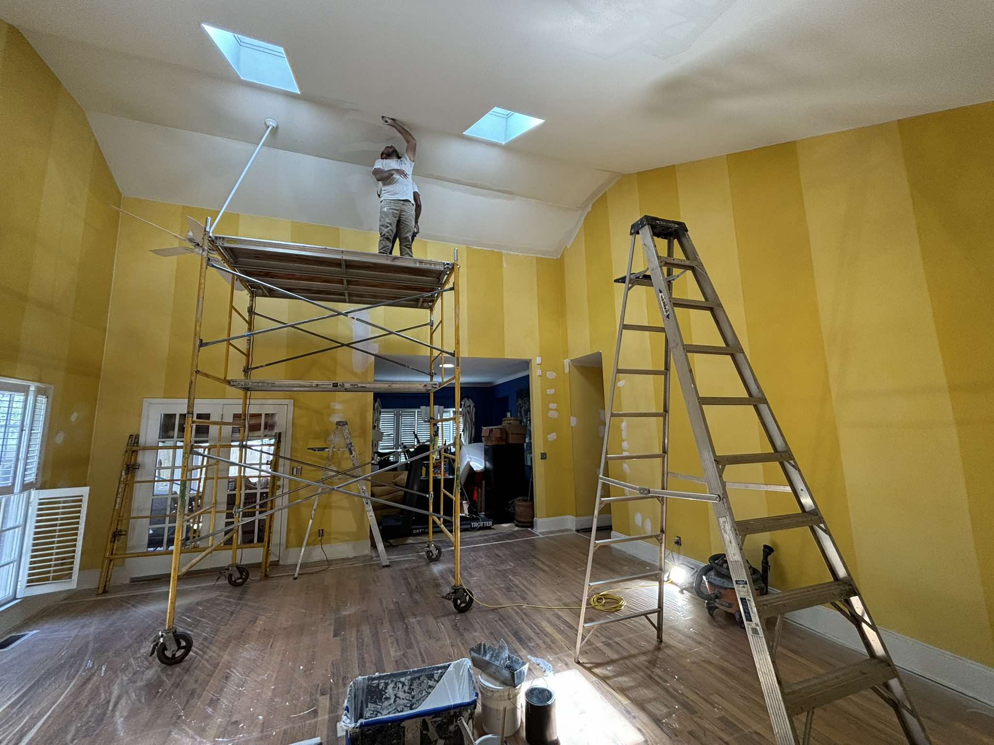 Habitación de techo alto durante la pintura con paredes amarillas, andamio móvil, escaleras y pisos de madera protegidos